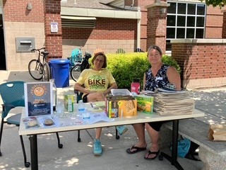 Two volunteers sit behind an outreach table displaying books, giveaways, and informational materials at an outdoor community event.