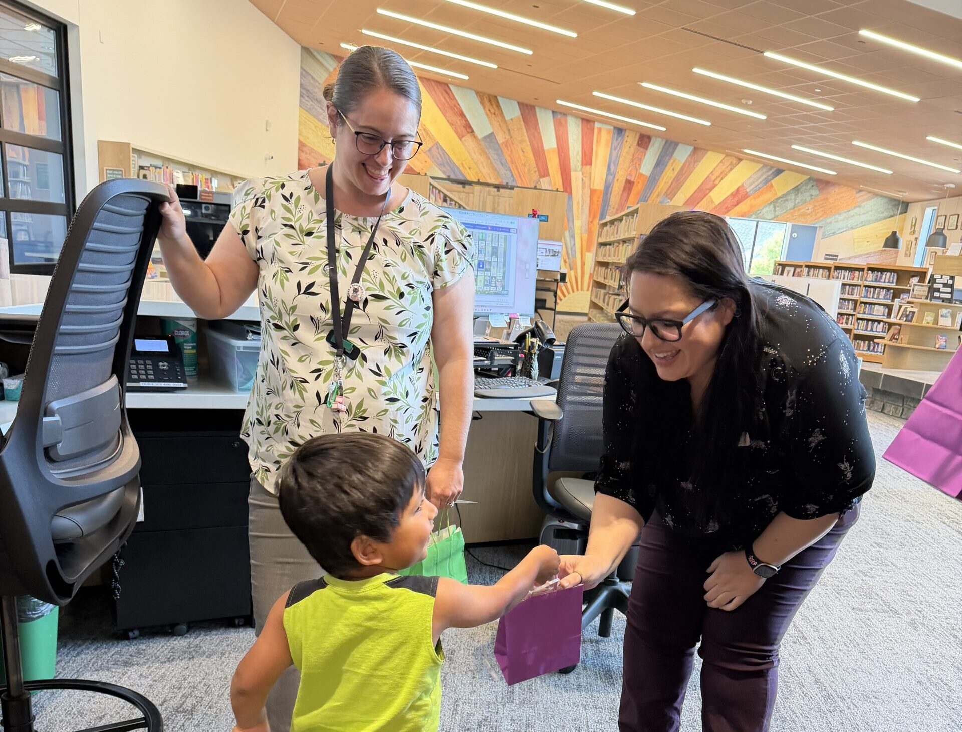 Two library staff members smile and interact with a young child. He is handing them gift bags.