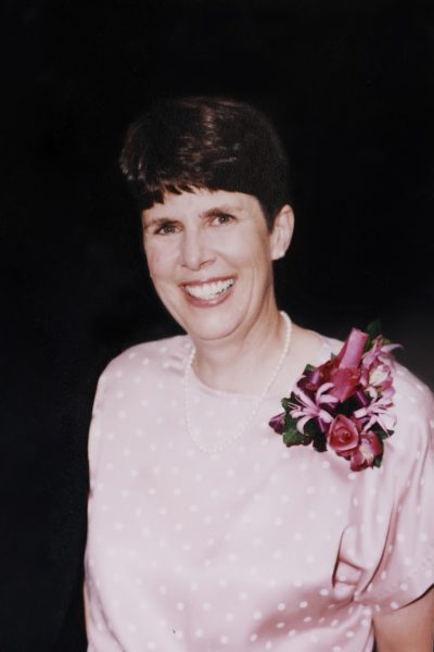 A smiling woman wearing a pink dress and floral corsage poses for a portrait against a dark background.